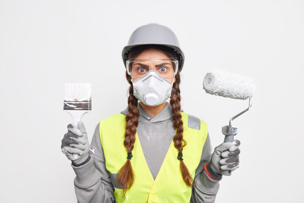 indoor shot of attentive busy woman builder with pigtails looks stunned through transparent safety glasses holds paint brush and roller involvd in reconstruction wears protective helmet and face mask